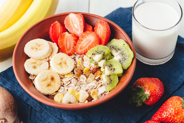 Cereal muesli with strawberries, banana and kiwi in the bowl, glass with milk on wooden white background, healthy breakfast