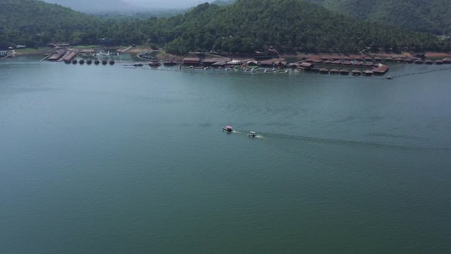 Wide orbit aerial shot above peaceful waters of Srinakarin dam. Lush, tropical jungle mountains in background with floating holiday resort cabins on the coastline as boats sail towards the village.