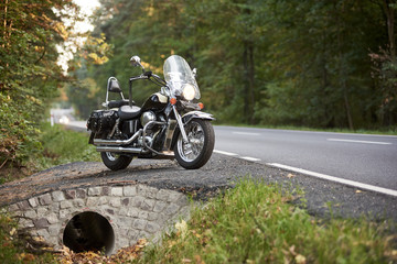 Black shiny powerful motorcycle parked on roadside on background of green trees on sunny summer day. Modern transportation technology, speed and long-distance traveling concept.