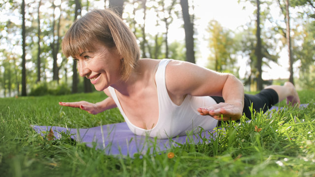 Photo of smiling happy woman 40 years old doing yoga exercises on fitness mat at forest. Harmony of human in nature. Middle aged people taking car of mental and physical health