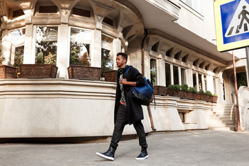Obraz premium A young man wearing in casual clothes, with backpack on shoulders, walking on street in day time, near buildings and sign crosswalk.