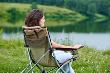 young woman freelancer sitting on chair and relaxing in nature near the lake. Outdoor activity in summer. Adventure traveling in national park. leisure, vacation, relaxation