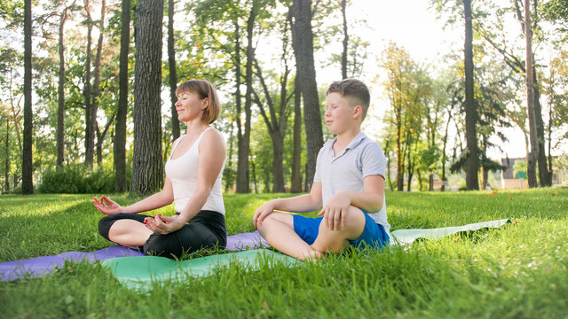 Photo of mother with her teenage boy son practising yoga asana on grass at park. Family doing fitness and sports outtodr at forest