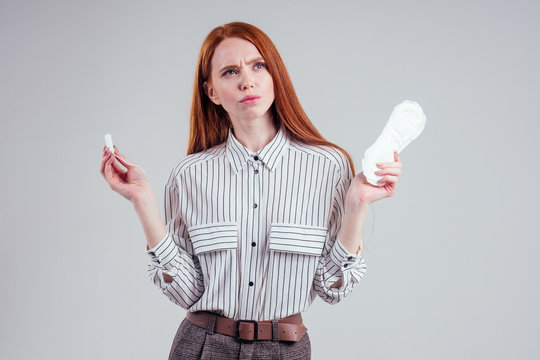 Closeup photo of young redhead businesswoman in striped shirt holding hygiene pad choosing tampon studio white background