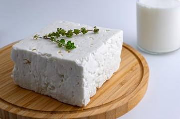 Large piece of traditional greek feta cheese on wooden cutting board with oregano and a bottle of milk on white background