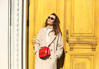 Trendy fashionable young female in a coat and red cross body bag against yellow background....