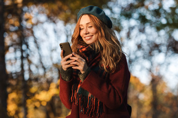 Happy young pretty woman walking outdoors in autumn spring park using mobile phone chatting.