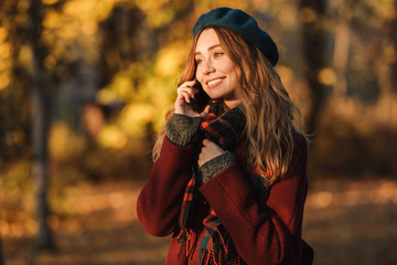 Beautiful happy young pretty woman walking outdoors in autumn spring park talking by mobile phone.