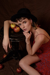 Cute young woman in vintage red dress and black hat with apples on brown fabric background. Conceptual fashion art photography