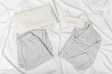 Grey pajamas, sleep mask, soft fluffy cushion on white crumpled sheet. Sleepwear for slumber on bed. Top view. Flat lay.