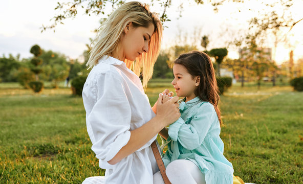 Side view image of happy child playing with pretty mother in the park. Beautiful young woman and her daughter relaxing on the green grass. Mom and little girl shares love. Happy Mothers Day.
