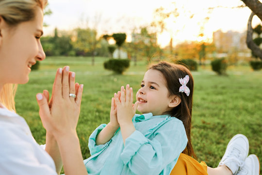 Horizontal outdoor image of cute little girl playing with her happy mother, touching hands by hands. Young woman and her daughter spending time together in the park. Mom and kid has fun. Mothers day