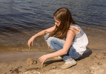 Fototapeta na wymiar girl on the river bank plays in the sand, builds a sand castle