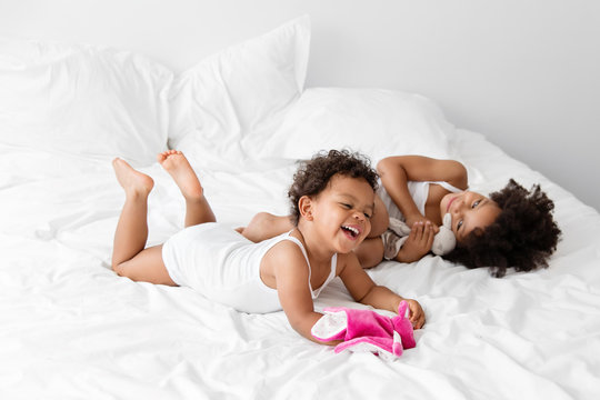 Two little girls having fun on white bed holding dolls