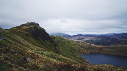 Stunning mountain view from the top seeing a lake, rugged valley and moody skies – captured during a hike at Snowdon in winter (Snowdonia National Park, Wales, United Kingdom)