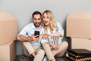 Photo of excited couple in casual clothing seating near cardboard boxes and using cellphone
