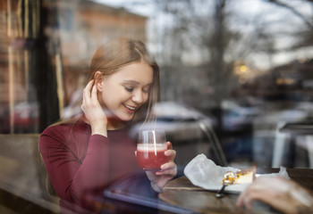 Pretty young woman with cocktail and cake at table in cafe, view from outdoors through window