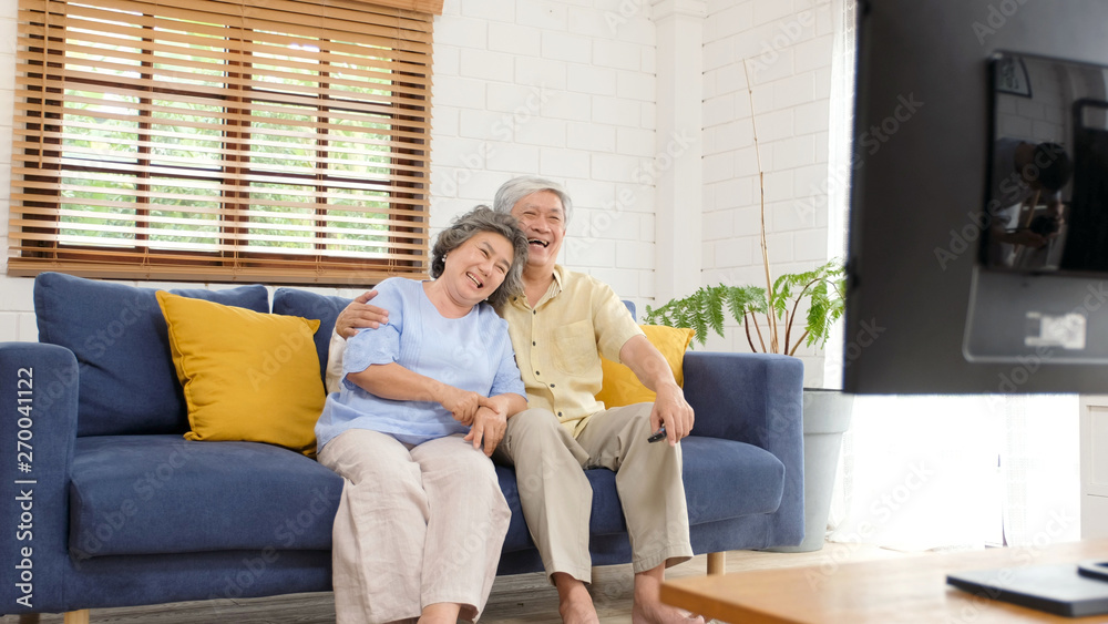 Wall mural Senior asian couple holding remote control while watching television in home living room with happiness, old people retirement routine lifestyle