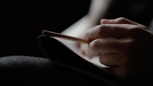 Close-up man's hand knocks nervously on the notebook with pencil on dark background. Person thinks at the cafe. Guy is considering a solution to the problem
