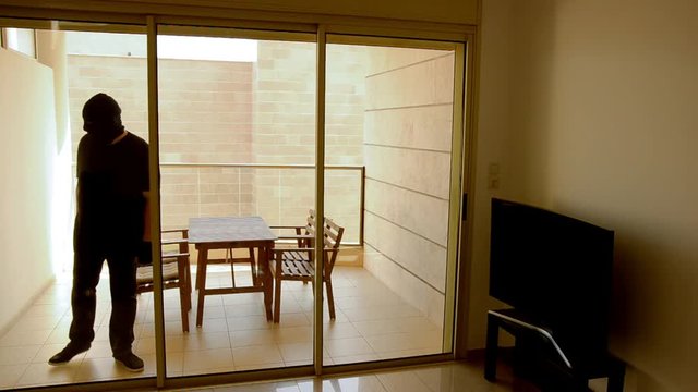 Thief in black balaclava gains access to the apartment through a balcony door.  Big build man has a screwdriver in his left hand. The man wearing black pants and a t-shirt with short sleeve.
