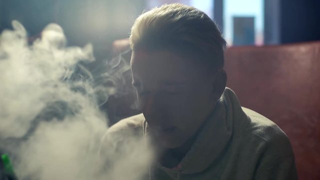Close-up handsome blonde guy with an earring in his ear smoking a hookah and exhales thick smoke at dark room of shisha lounge in slow motion. Young hipster man relaxing with his friends