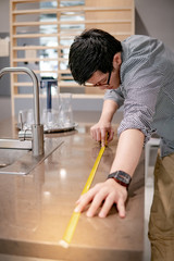Young Asian man using tape measure for measuring granite countertops on modern kitchen counter in showroom. Shopping furniture for home improvement. Interior design concept