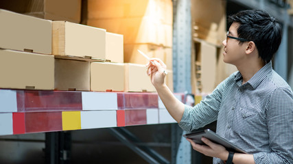 Young Asian man worker doing stocktaking of product in cardboard box on shelves in warehouse by using digital tablet and pen. Physical inventory count concept