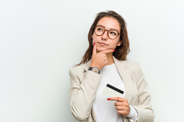 Young european business woman holding a credit card looking sideways with doubtful and skeptical expression.