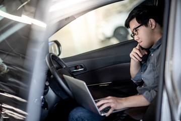 Overworked Asian businessman with glasses feeling stressed and tired while working with laptop computer on driver seat in the car. Male entrepreneur working hard for his business.