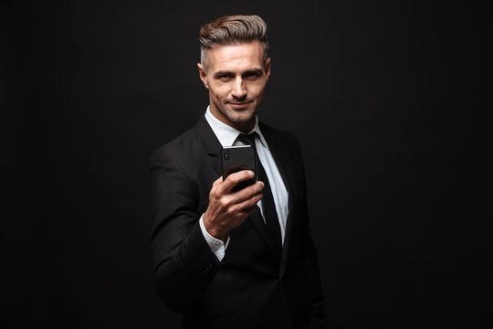 Portrait of adult unshaven businessman dressed in formal suit using cellphone and looking at camera