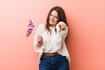 Obraz na płótnie Canvas Young plus size curvy woman holding a united kingdom flag cheerful smiles pointing to front.