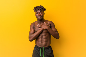 Young black man shirtless wearing swimsuit has friendly expression, pressing palm to chest. Love...