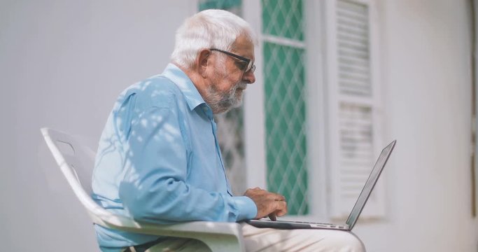 elderly man uses a laptop sitting on a chair in his backyard