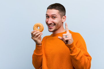 Young caucasian man holding a donut showing number one with finger.