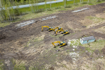 Aerial side view of four yellow crawler excavators standing on ground near the construction site and waiting for the working day to start digging the ground near the building, road in field among tree