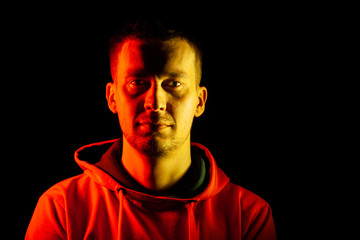 An adult guy stands look forward in an orange hooded sweatshirt highlighted in yellow and red on the sides with a smile divided in half and in the middle hidden by shadow on black isolated background