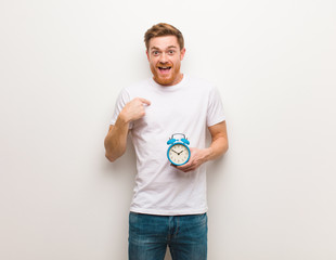 Young redhead man surprised, feels successful and prosperous. He is holding an alarm clock.