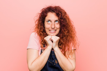 Young pretty ginger redhead woman wearing a jeans dungaree keeps hands under chin, is looking happily aside.