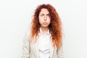 Young natural redhead business woman isolated against white background blows cheeks, has tired expression. Facial expression concept.