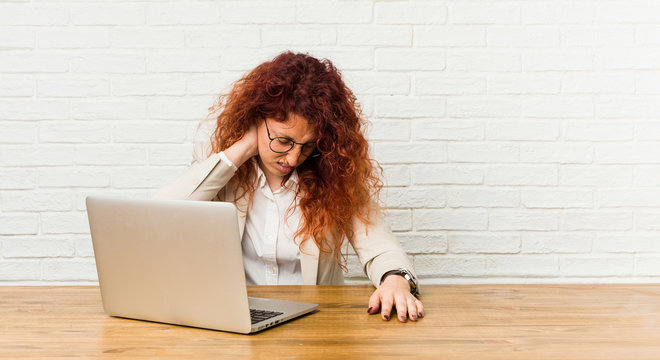 Young redhead curly woman working with her laptop suffering neck pain due to sedentary lifestyle.