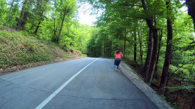 Wide forward tracking shot of an overweight woman wearing a long pony tail running on the road through a forest 