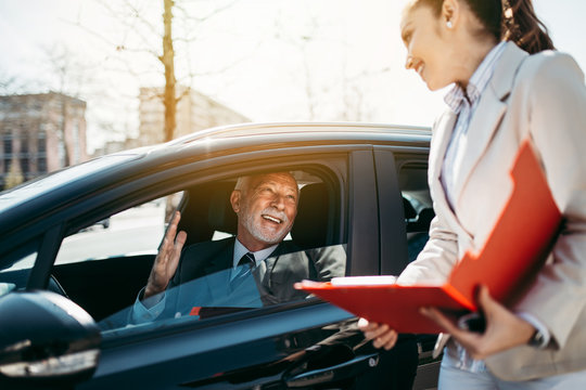 Beautiful Young Woman Car Dealer Or Sales Manager Showing Vehicle Specification To Happy Senior Man Buyer.