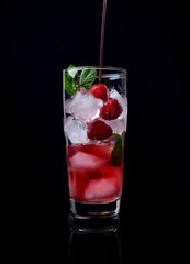 Process of pouring the red drink into a tall glass filled with ice cubes, raspberries and mint against the black background