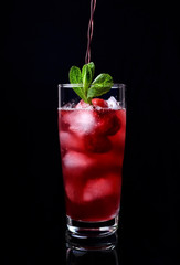 Process of pouring the red drink into a tall glass filled with ice cubes, raspberries and mint against the black background