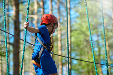 Young child boy in safety harness and helmet attached with carbine to cable moves confidently along rope way in recreation park.  Sport, game, leisure concept.