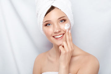 attractive young woman in bath towel applying cream on her face smiling and looking to the camera, girl doing face treatment