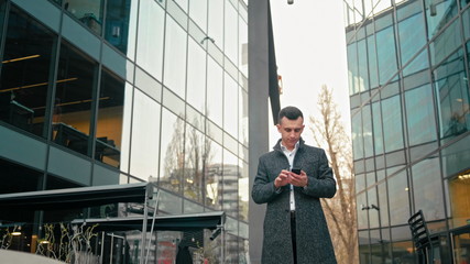 Adult Caucasian Confident Young Business Man is Using Smartphone App and Walking near Modern Glass Office Building in the City. Medium Long Low Angle