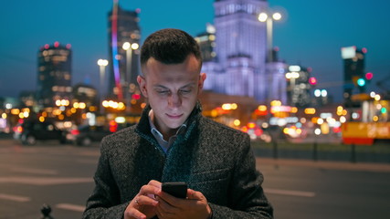 Adult Caucasian Confident Young Business Man Entrepreneur is Using Smartphone App Outdoors in the City in the Late Evening or Night with Beautiful Blurred Cityscape Lights