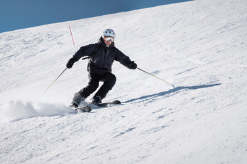 Fototapeta na wymiar Bearded male skier in a helmet and a ski mask rides on a slope at speed and brakes with snow powder on skis against the background of a ski slope and blue sky. Athlete in a black suit