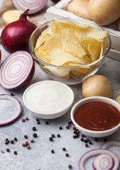 Fresh organic homemade potato crisps chips in glass bowl with sour cream and red onions on light kitchen table background. Bowl of ketchup and green onion
